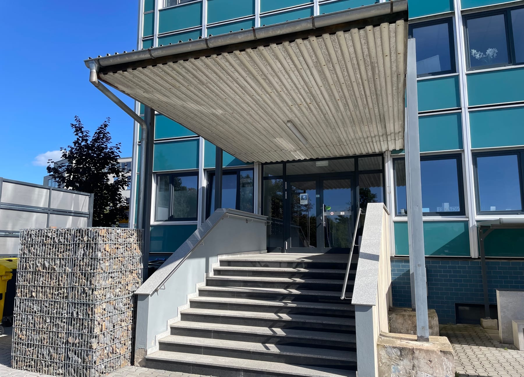 Hintereingang des Gebäudes: eine Treppe mit sieben Stufen und beidseitigem Handlauf führt unter einem Vordach zur Eingangstür